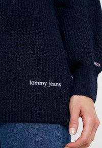 Granatowy sweter z dzianiny z ribbowym wzorem, okrągłym dekoltem i białym haftowanym logo "tommy jeans" z przodu. Widoczne wykończenia mankietów.