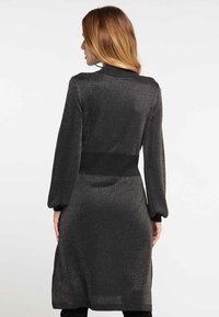 Femme avec de longs cheveux châtains clairs portant une robe noire à manches longues scintillante avec un détail de taille cintrée, vue de dos sur fond uni.