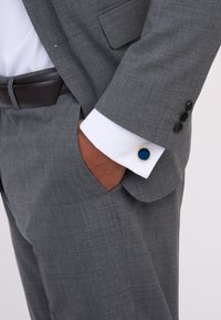 Grauanzug aus strukturiertem Stoff, kombiniert mit einem kontrastierenden weißen Hemd, braunem Gürtel und einem Manschettenaufschlag mit blauen und goldenen Akzenten.