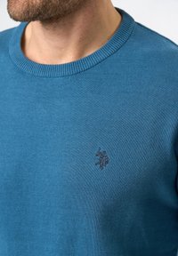 Niebieski sweter dzianinowy z okrągłym dekoltem. Na piersi subtelne haftowane logo. Gładka faktura i prążkowany kołnierz.