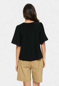 Top nero a maniche corte con design fluido e schiena plissettata, realizzato in tessuto leggero e con scollatura rotonda, abbinato a pantaloni beige.