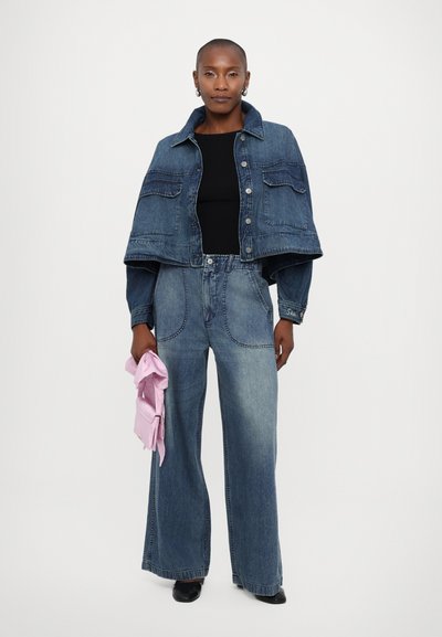 Jeans jakna s skrajno zasnovo, dvema sprednjima žepoma, v kombinaciji z širokimi denim hlačami. Model drži svetlo roza torbico.