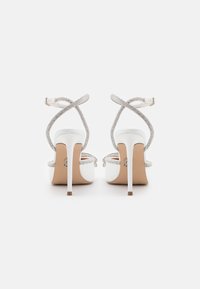 White high-heeled sandals with crisscross straps adorned with silver rhinestones. Features a smooth finish and a slender stiletto heel.