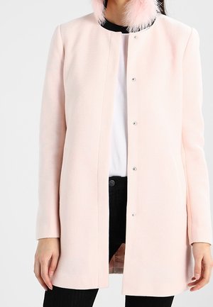 Woman wearing a pale pink textured long coat with snap buttons, paired with a white shirt and black pants.