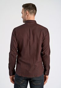 Camisa de manga larga color granate con cuello abotonado, canesú trasero recto y puños abotonados. Se lleva con vaqueros oscuros, mostrando una textura suave.