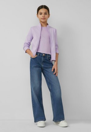 Junges Mädchen steht mit einer Hand in der Tasche, trägt eine lavendelfarbene Zip-up-Jacke, passendes Oberteil, weite blaue Jeans und weiße Turnschuhe vor einer grauen Wand.