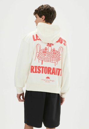 Junger Mann trägt einen weißen Hoodie mit rotem, restaurantbezogenem Grafik- und Textdruck auf dem Rücken, kombiniert mit schwarzen Shorts, seitlich stehend.