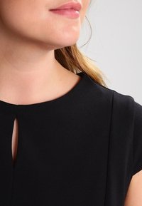 Detailní záběr dolní části obličeje a krku ženy, která má na sobě černý top s malým předním rozparkem u výstřihu.