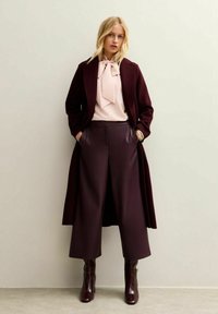 Burgundy long coat over a pink blouse with a bow detail, paired with wide-leg trousers and knee-high dark boots. Sleek textures and tailored fit.