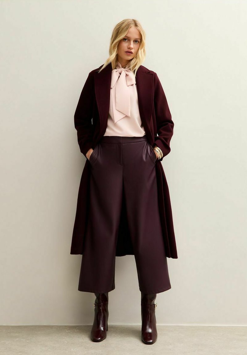 Burgundy long coat over a pink blouse with a bow detail, paired with wide-leg trousers and knee-high dark boots. Sleek textures and tailored fit.
