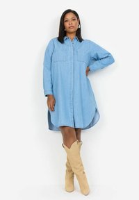 Gaiši zils džinsa krekla kleita ar garām piedurknēm, pogām priekšpusē, divām krūšu kabatām un izliekto apakšmalu, savienota ar ceļgaliem augstiem bēšiem zābakiem.