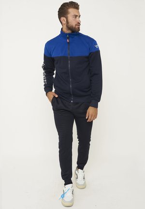 Uomo con giacca blu e nera con cerniera e pantaloni neri in piedi con le mani in tasca, che guarda verso destra, indossa scarpe sportive bianche.