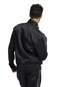 Svart satinjacka med hög krage, dragkedjestängning och vita sidostråkar. Mjuk textur med elastiska muddar och fåll.