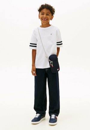 Lächelnder Junge, der ein weißes T-Shirt mit schwarzen Streifen an den Ärmeln, dunkle, weit geschnittene Jeans, marineblaue Sneakers und eine kleine navyblaue Umhängetasche trägt.