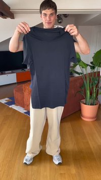 Giovane uomo in piedi nel soggiorno che tiene in mano una maglietta nera semplice, indossando pantaloni beige e sneakers bianche, con una pianta e un divano nelle vicinanze.