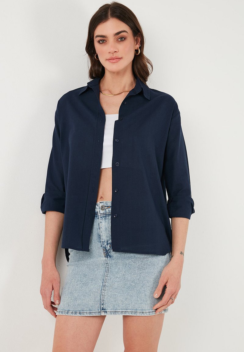 Camicia blu navy con bottoni in tessuto leggero, con maniche arrotolate, abbinata a una mini gonna di jeans chiari. Design semplice e classico, senza motivi.