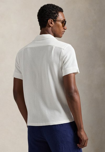 Λευκό πλεκτό πόλο πουκάμισο με γιακά και κοντά μανίκια, με υφή ύφανσης. Το πουκάμισο συνδυάζεται με παντελόνι σε ναυτικό μπλε χρώμα.