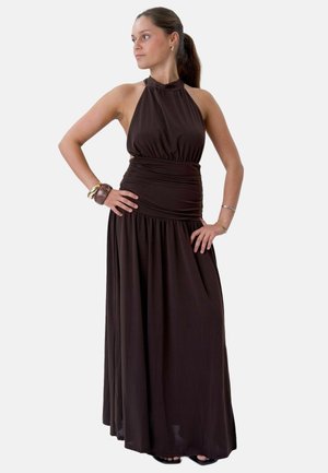 Femme debout les mains sur les hanches, portant une robe longue sans manches marron foncé à col halter et des bracelets, regardant vers sa gauche.