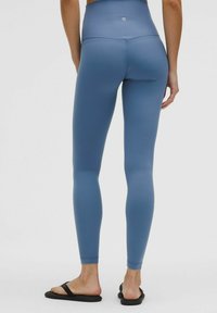 Magas derekú kék leggings rugalmas anyagból. Sima felülettel és testhezálló kialakítással rendelkezik. Fekete papucsokkal viselik.