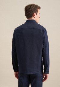 Camicia blu navy con maniche lunghe, polsini a bottoni e orlo curvo. Texture liscia con un design minimalista e dettagli delle cuciture sottili.