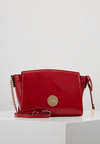 Borsa a tracolla in pelle rossa con catena dorata e piatto logo circolare oro su una superficie bianca contro uno sfondo grigio chiaro.