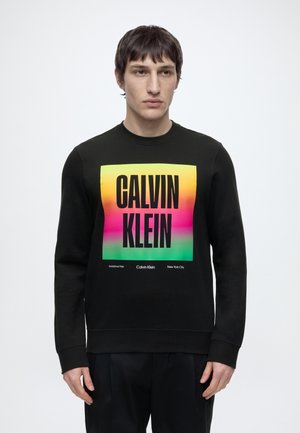 Ung mand iført sort sweatshirt med stort farverigt Calvin Klein logo firkant foran, stående mod en ensfarvet lys baggrund.
