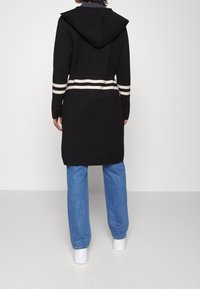 Schwarze Kapuzenjacke mit weißen Streifen an der Taille und an den Ärmeln. Lange Ärmel, lockere Passform und ein schlanker Saum. Getragen über blauen Jeans und weißen Turnschuhen.