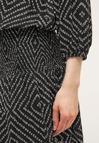 Černobílé šaty s geometrickým potiskem, s nařaseným pasem a nabíranými rukávy, vyrobené z lehkého materiálu.