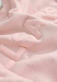 Felpa rosa chiaro con testo in rilievo e una superficie morbida e texturizzata. Il tessuto sembra lavorato a maglia e ha una sensazione di comfort.