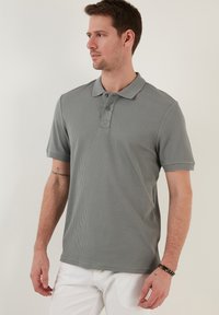 Harmaa ribbattu polo-paita lyhyillä hihoilla, kolmen napin nappilista ja hienovarainen ompeluyksityiskohta rinnassa. Yhdistetty valkoisiin housuihin.