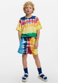 T-shirt tie-dye in giallo, blu, rosso e verde con logo "Desigual"; abbinato a pantaloni corti tie-dye coordinati e calze blu fin sopra il ginocchio.