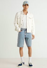 Veste en denim blanche, t-shirt blanc, shorts en denim bleu clair et baskets navy avec des accents blancs. Le mannequin porte une casquette grise claire.