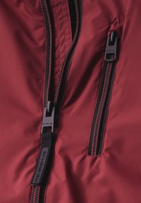 Tissu de veste bordeaux avec deux zips noirs, une poche avec étiquette et un détail de couture contrasté. Texture lisse avec un design moderne.