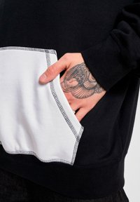 Zwarte sweatshirt met een grote witte zak. Heeft contrasterende stiksels en een zachte textuur. Hand zichtbaar binnen de zak.