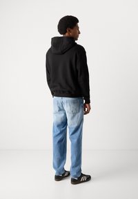 Felpa nera con cappuccio e cordino, jeans sbiaditi blu chiaro e scarpe nere con strisce bianche. Soggetto visto da dietro, che mostra un outfit casual.
