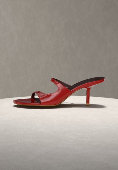 Červená lodička z lakovanej kože so zašitou špičkou, ohnutým remienkom a tenkým stredne vysokým podpätkom na neutrálnom povrchu a pozadí.