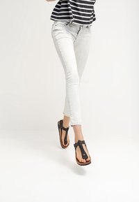 Chanclas de goma negra con diseño de tira para los dedos, correa ajustable y suela marrón texturizada. Combinadas con unos vaqueros ajustados de color claro.