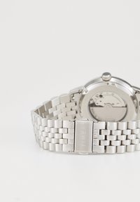 Silberne Timex-Armbanduhr aus Metall mit einem Gliederarmband und sichtbarem mechanischem Uhrwerk auf der Rückseite des Zifferblatts.