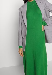 Robe en maille côtelée verte avec un col montant, associée à un blazer gris clair. La robe présente des manches longues et une silhouette ajustée.