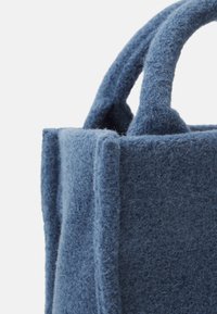 Blå filt tote bag med teksturert overflate, med runde håndtak og vinklete sider. Ren, minimalistisk design uten synlig maskinvare.