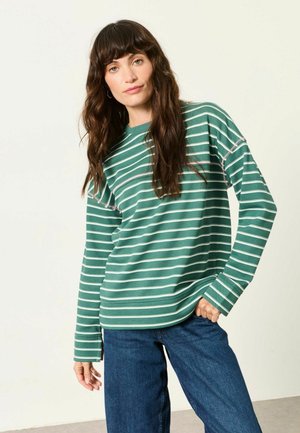 Kvinde med langt brunt hår iført en grøn og hvid stribet langærmet skjorte og blå jeans, der står mod en ensfarvet lys baggrund.