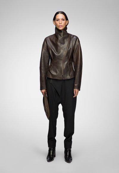 Brown leather jacket with a high collar, asymmetrical zipper, and smooth texture, paired with loose black pants and ankle boots.