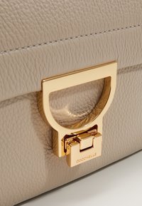 Sac à main en cuir beige texturé avec un fermoir rectangulaire doré comportant une poignée semi-circulaire et une gravure de marque.
