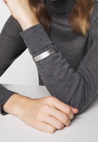 Bracelet en métal argenté avec un motif carré, porté sur un poignet vêtu d'un pull gris, présentant un design moderne et une finition texturée.