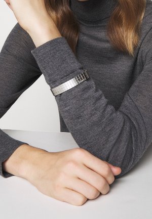 Bracelet en métal argenté avec un motif carré, porté sur un poignet vêtu d'un pull gris, présentant un design moderne et une finition texturée.