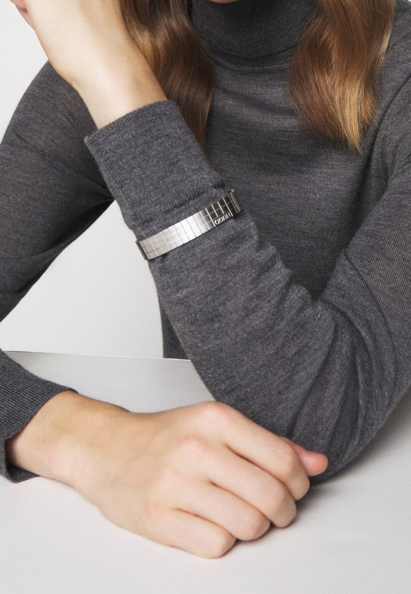 Bracelet en métal argenté avec un motif carré, porté sur un poignet vêtu d'un pull gris, présentant un design moderne et une finition texturée.