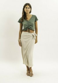 Haut vert court avec un décolleté en V et un devant froncé, associé à une jupe portefeuille beige arrivant au genou avec un détail de nœud sur le côté. Sandales à lanières.