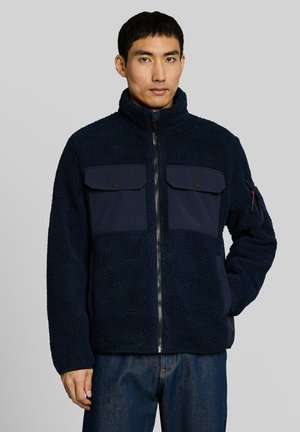 Uomo che indossa una giacca in pile blu navy con due tasche sul petto e una cerniera, in piedi con una mano nella tasca contro uno sfondo grigio uniforme.