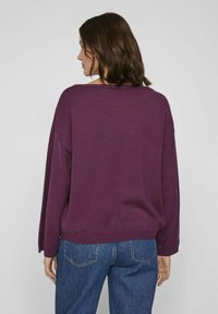 Pull en tricot violet avec une coupe ample, un grand col et des manches longues. Associé à un jean bleu, mettant en avant un style décontracté et informel.