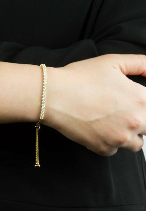 Håndled med et gyldent og diamant tennisarmbånd med justerbar kæde, der hænger, mod en sort langærmet beklædning.